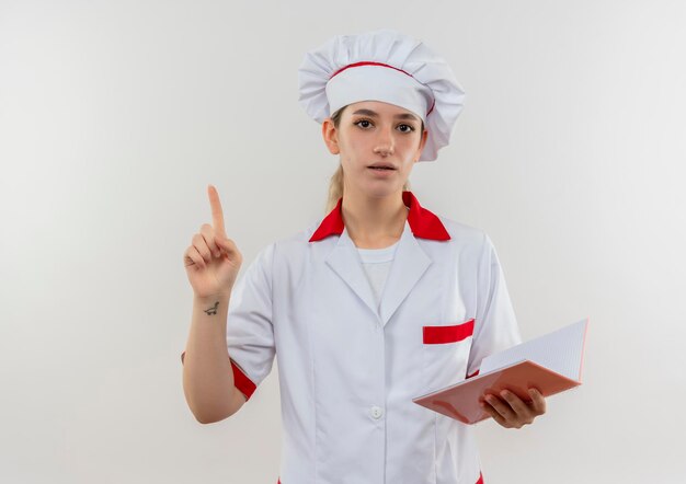 Jeune jolie cuisinière impressionnée en uniforme de chef tenant un bloc-notes et levant le doigt isolé sur un mur blanc