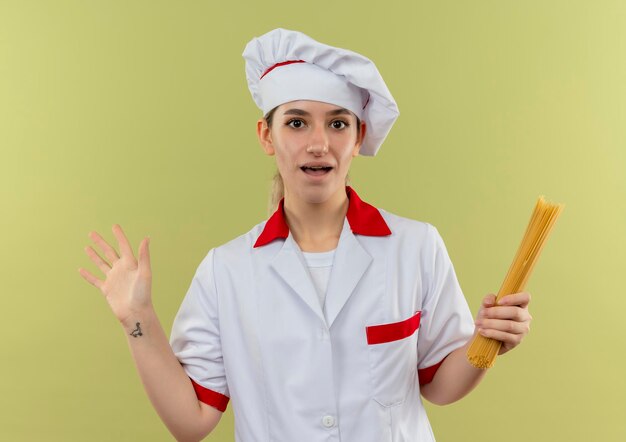 Jeune jolie cuisinière impressionnée en uniforme de chef tenant des pâtes spaghetti montrant une main vide isolée sur un mur vert