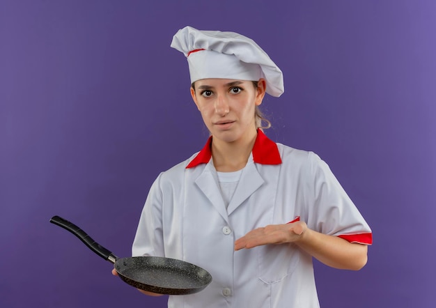 Jeune jolie cuisinière impressionnée en uniforme de chef tenant et pointant avec la main une poêle à frire isolée sur un mur violet