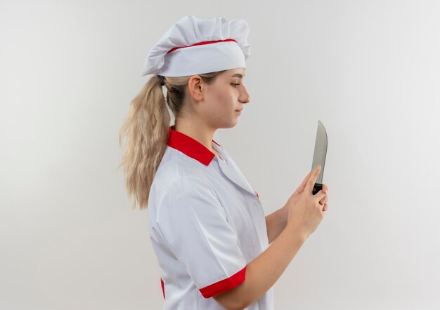 Jeune jolie cuisinière en uniforme de chef debout en vue de profil tenant et regardant le couteau