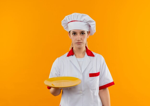 Jeune jolie cuisinière en uniforme de chef tenant une assiette vide à la recherche