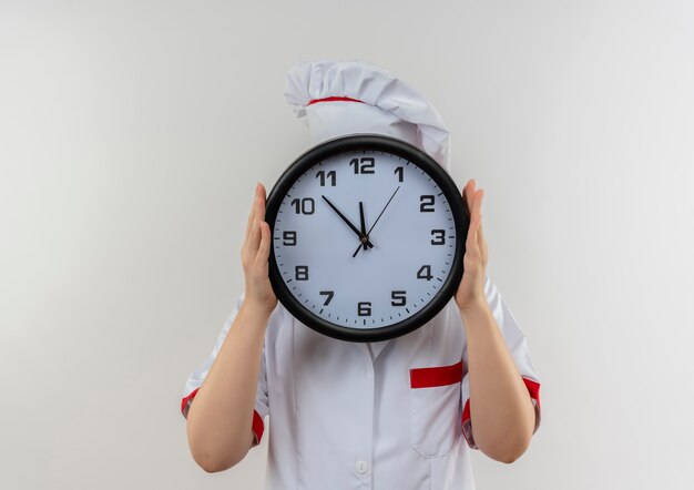 Jeune jolie cuisinière en uniforme de chef tenant horloge et se cachant derrière elle