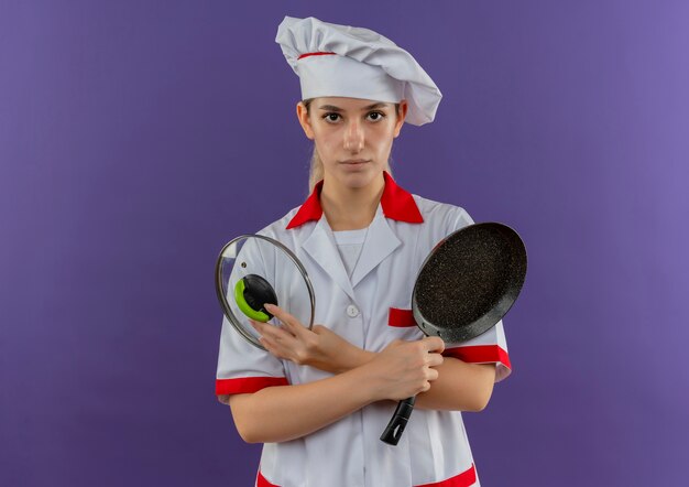 Jeune jolie cuisinière en uniforme de chef tenant une poêle et un couvercle de casserole à