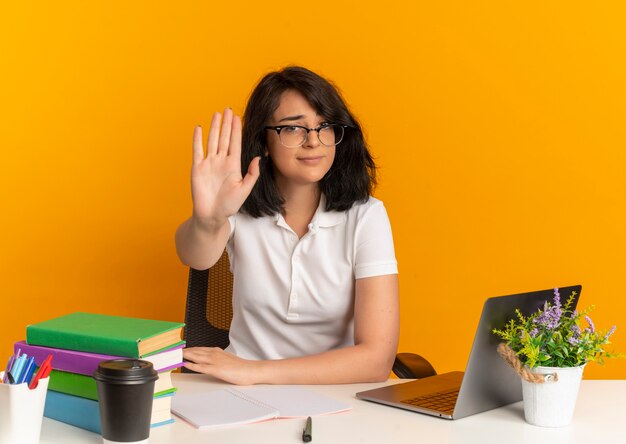 Jeune jolie écolière caucasienne confiante portant des lunettes est assise au bureau avec des gestes d'outils scolaires s'arrêtent sur orange avec copie espace