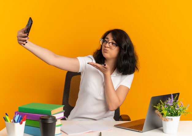 Jeune jolie écolière caucasienne confiante portant des lunettes est assise au bureau avec des outils scolaires regarde l'envoi de téléphone baiser avec la main sur l'orange avec copie espace