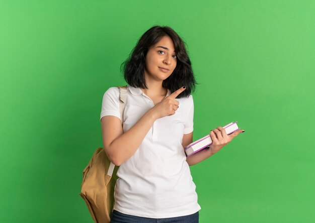 Jeune jolie écolière caucasienne confiante portant des points de sac à dos sur le côté tenant des livres sur vert avec copie