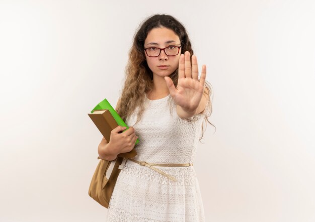 Jeune jolie écolière portant des lunettes et sac à dos tenant des livres à la recherche et aux gestes arrêt à la caméra isolé sur fond blanc avec espace de copie