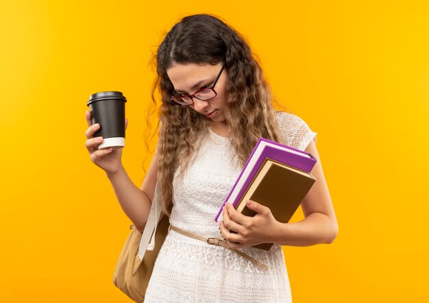 Jeune jolie écolière portant des lunettes et sac à dos tenant une tasse de café en plastique et des livres regardant vers le bas isolé sur fond jaune avec espace de copie