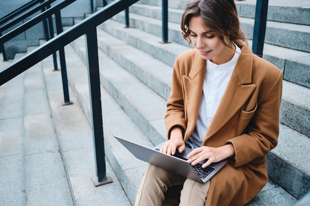 Jeune jolie femme d'affaires en manteau travaillant rêveusement avec un ordinateur portable dans les escaliers en plein air