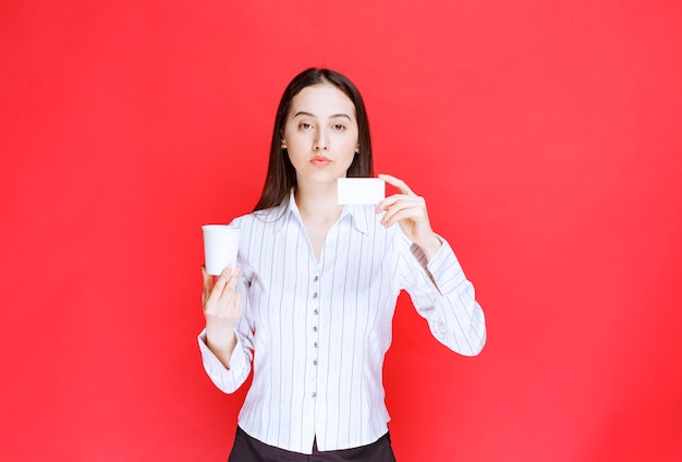 Jeune jolie femme d'affaires tenant une tasse en plastique et une carte de visite sur fond rouge.