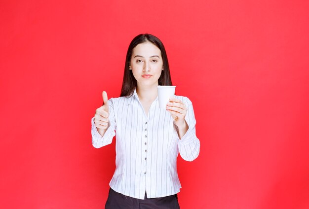 Jeune jolie femme d'affaires tenant une tasse en plastique et donnant le pouce vers le haut.