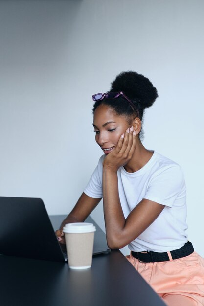 Jeune jolie femme afro-américaine décontractée travaillant avec plaisir sur un ordinateur portable avec du café pour aller dans un bureau moderne