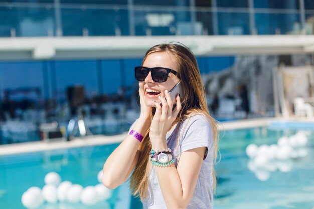 Jeune jolie femme aux cheveux longs et lunettes de soleil noires se tient près de la piscine