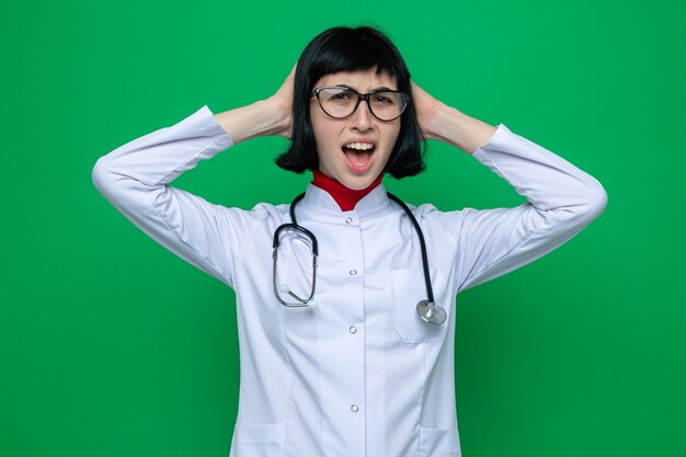 Jeune jolie femme caucasienne agacée avec des lunettes en uniforme de médecin avec stéthoscope met les mains sur la tête