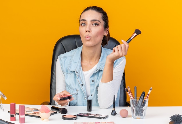 Jeune jolie femme caucasienne assise à table avec des outils de maquillage faisant un visage de poisson tenant un blush et un pinceau de maquillage isolé sur un mur orange avec espace pour copie