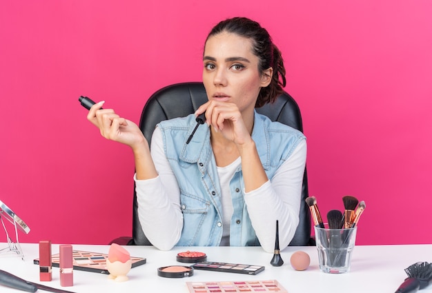 Jeune jolie femme caucasienne assise à table avec des outils de maquillage tenant du mascara isolé sur un mur rose avec espace de copie