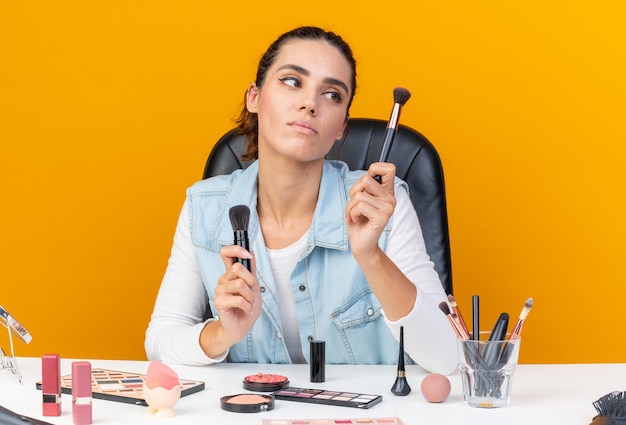 Jeune Jolie Femme Caucasienne Assise à Table Avec Des Outils De Maquillage Tenant Et Regardant Des Pinceaux De Maquillage