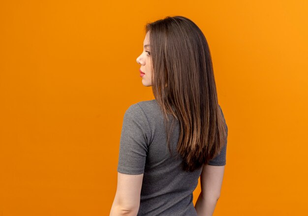 Jeune jolie femme debout en vue de derrière et regardant à côté isolé sur fond orange avec copie espace