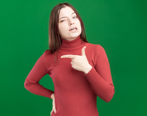 Jeune jolie femme douteuse gardant la main sur la taille pointant sur le côté isolé sur un mur vert avec espace pour copie