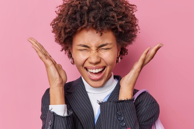 Jeune jolie femme émotionnelle positive aux cheveux bouclés lève les paumes près de gead s'exclame fort garde les yeux fermés porte une veste rayée noire réagit sur de merveilleuses nouvelles isolées sur fond rose.