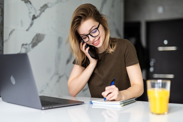 Jeune jolie femme est assise à la table de la cuisine à l'aide d'un ordinateur portable et parle sur un téléphone portable