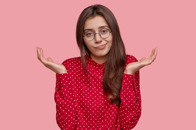 Jeune jolie femme étant à perte, montre un geste impuissant, ne peut pas prendre de décision, écarte les mains, porte des lunettes optiques