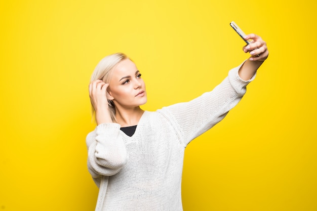 Jeune jolie femme femme en pull blanc fait selfie sur son smartphone sur jaune