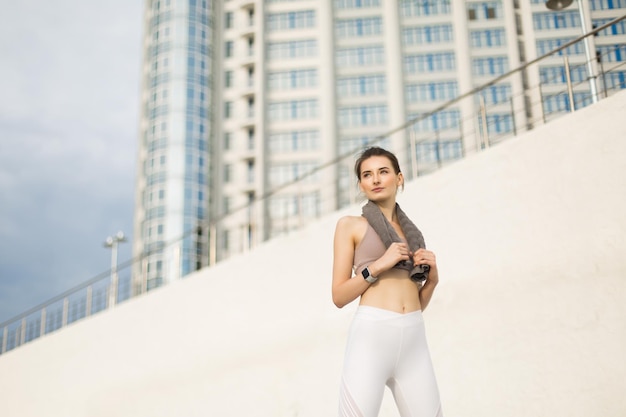 Jeune jolie femme en haut sportif et leggings blancs tenant une petite serviette sur les épaules tout en regardant rêveusement de côté sur fond flou