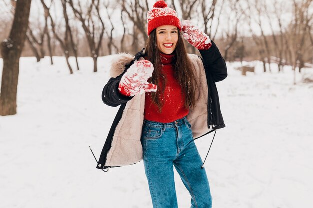 Jeune jolie femme heureuse souriante dans des mitaines rouges et bonnet tricoté portant manteau d'hiver marchant dans le parc dans la neige, des vêtements chauds, s'amuser