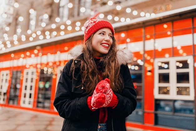 Photo gratuite jeune jolie femme heureuse souriante dans des mitaines rouges et bonnet tricoté portant un manteau d'hiver marchant dans la rue de la ville, des vêtements chauds