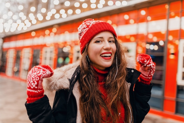 Jeune jolie femme heureuse souriante dans des mitaines rouges et bonnet tricoté portant un manteau d'hiver marchant dans la rue de la ville, des vêtements chauds