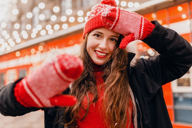 Jeune jolie femme heureuse souriante dans des mitaines rouges et bonnet tricoté portant un manteau d'hiver marchant dans la rue de la ville, des vêtements chauds