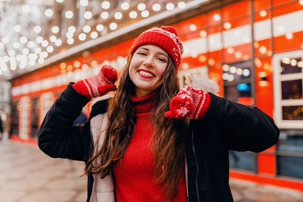 Jeune jolie femme heureuse souriante dans des mitaines rouges et bonnet tricoté portant un manteau d'hiver marchant dans la rue de la ville, des vêtements chauds