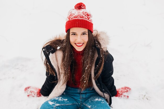 Jeune jolie femme heureuse souriante en mitaines rouges et bonnet tricoté portant un manteau d'hiver assis sur la neige dans le parc, des vêtements chauds