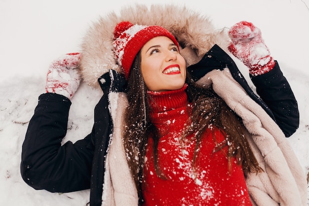 Jeune jolie femme heureuse souriante en mitaines rouges et bonnet tricoté portant manteau d'hiver couché dans le parc dans la neige, des vêtements chauds, vue d'en haut