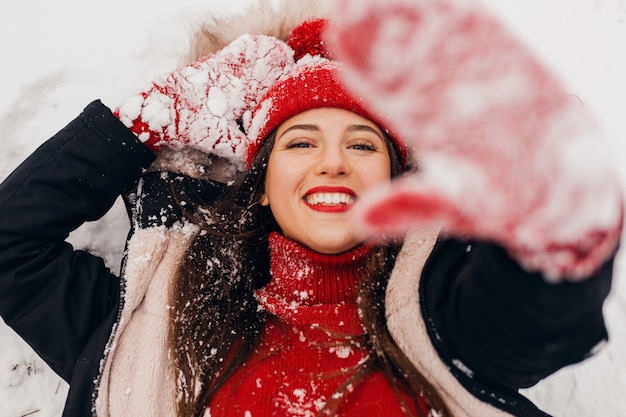 Jeune jolie femme heureuse souriante en mitaines rouges et bonnet tricoté portant manteau d'hiver couché dans le parc dans la neige, des vêtements chauds, vue d'en haut