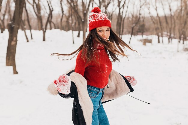 Jeune jolie femme heureuse souriante en mitaines rouges et bonnet tricoté portant un manteau d'hiver, marchant dans le parc dans la neige, des vêtements chauds