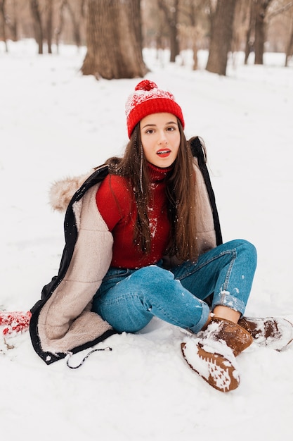 Jeune jolie femme heureuse souriante en mitaines rouges et bonnet tricoté portant un manteau d'hiver, marchant dans le parc, jouant avec la neige dans des vêtements chauds