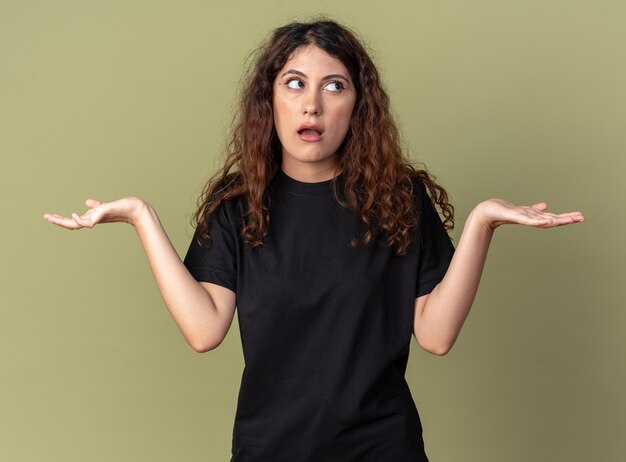 Jeune jolie femme ignorante regardant le côté montrant les mains vides isolées sur le mur vert olive