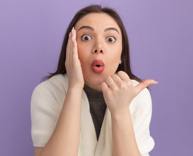 Jeune jolie femme impressionnée gardant la main près de la bouche pointant sur le côté chuchotant isolé sur un mur violet