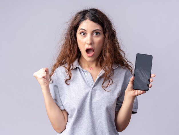 Jeune jolie femme impressionnée regardant à l'avant montrant un téléphone portable à l'avant faisant un geste oui isolé sur un mur blanc