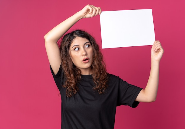 Photo gratuite jeune jolie femme impressionnée tenant du papier blanc au-dessus de la tête en le regardant isolé sur un mur cramoisi