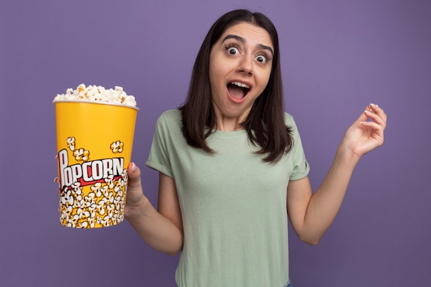 Jeune jolie femme impressionnée tenant un seau de pop-corn et un morceau de pop-corn regardant à l'avant isolé sur un mur violet