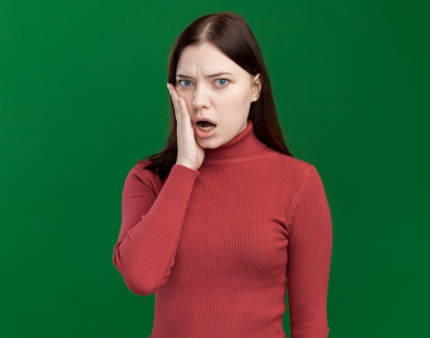 Jeune jolie femme inquiète regardant l'avant en gardant la main sur le visage isolé sur un mur vert avec espace pour copie