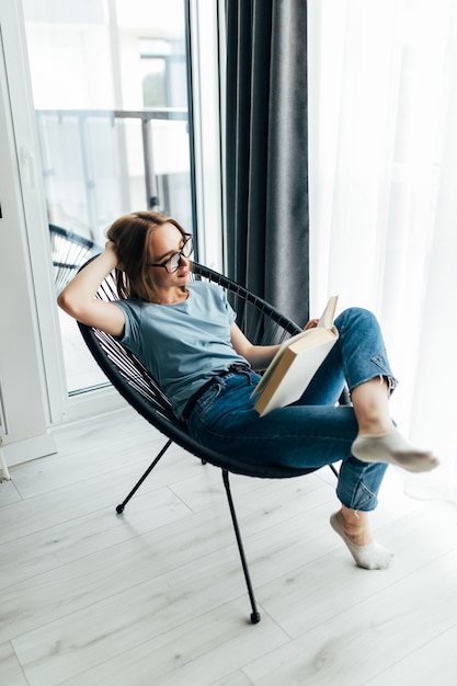 Jeune jolie femme lisant un livre et assise sur une chaise confortable à la maison