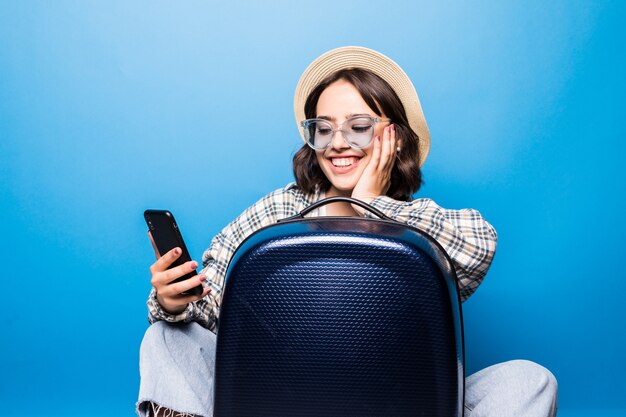 Jeune jolie femme à lunettes de soleil et chapeau de paille avec une valise se penche sur le téléphone isolé. Voyage en vol.