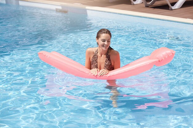 Jeune jolie femme avec matelas gonflable rose flottant dans la piscine