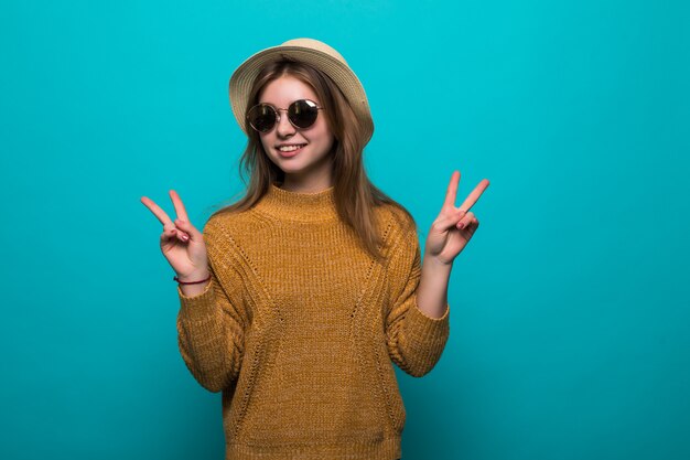 Jeune jolie femme portant chapeau et lunettes de soleil a souligné le geste de paix sur le mur bleu