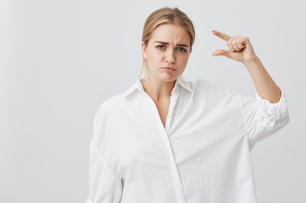 Jeune jolie femme portant une chemise blanche montrant quelque chose de très peu avec les mains tout en gesticulant. Étudiante blonde démontrant la taille de quelque chose