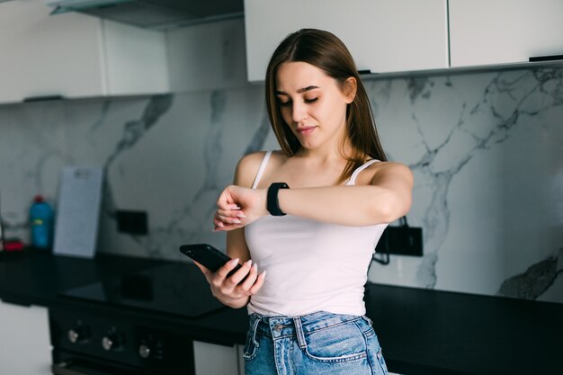 Jeune jolie femme prenant à sa montre-bracelet alors qu'il était assis sur une table dans une cuisine